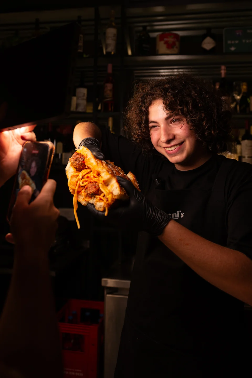 Persona con guantes negros presentando un sándwich hacia una cámara