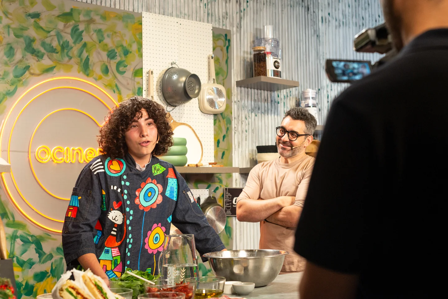 Dos personas junto a una mesada de cocina en un estudio con cartelería cocineros