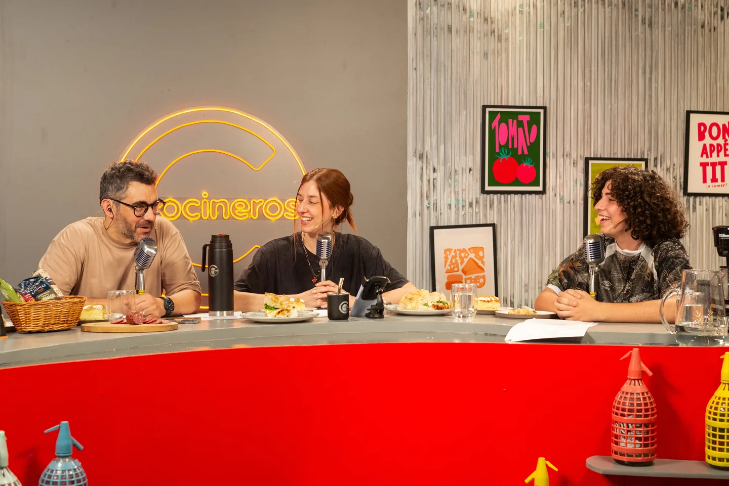 Tres personas conversando en torno a una mesa con alimentos y micrófonos en un estudio