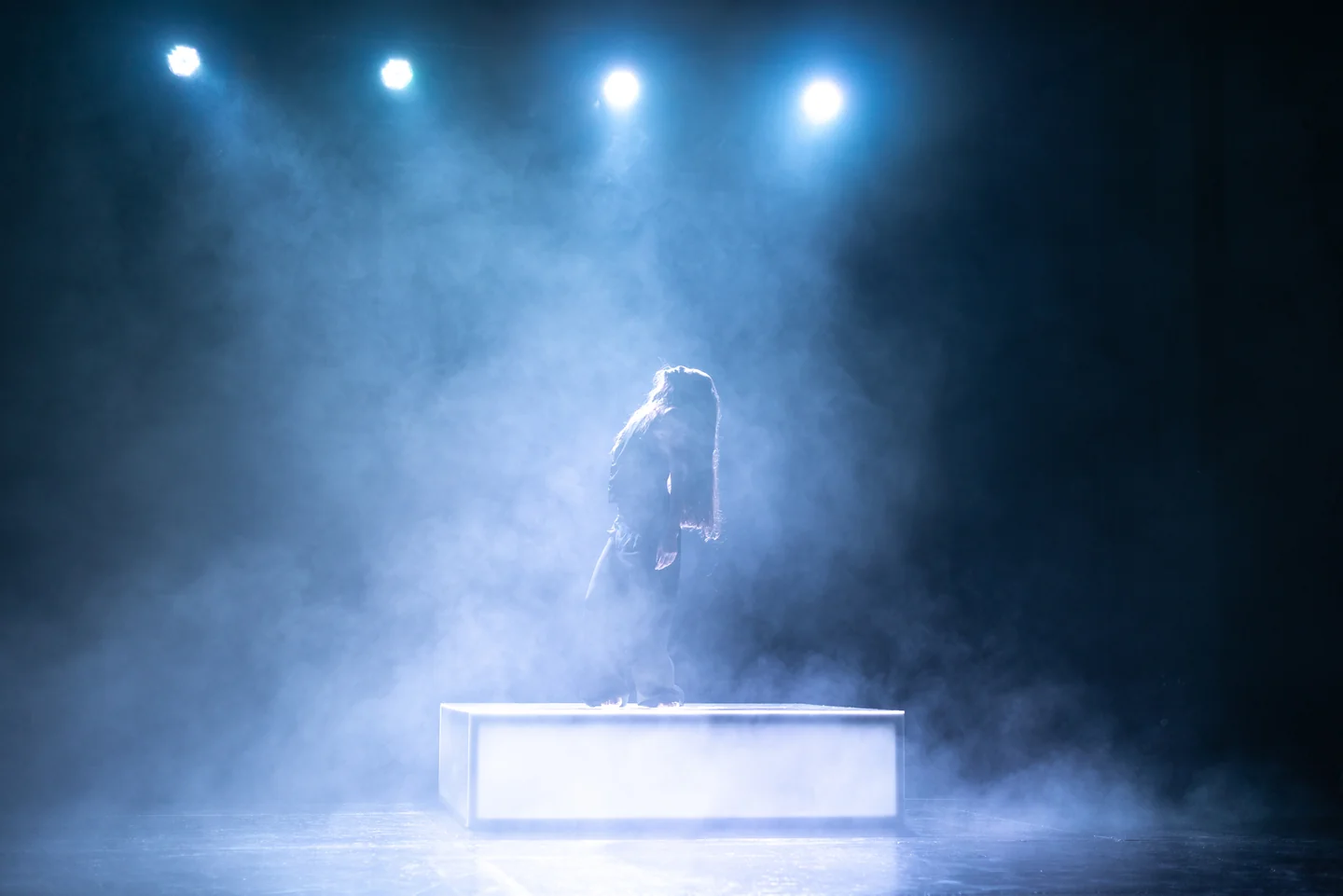 Figura sobre una plataforma luminosa en un escenario con humo azul y cuatro focos superiores