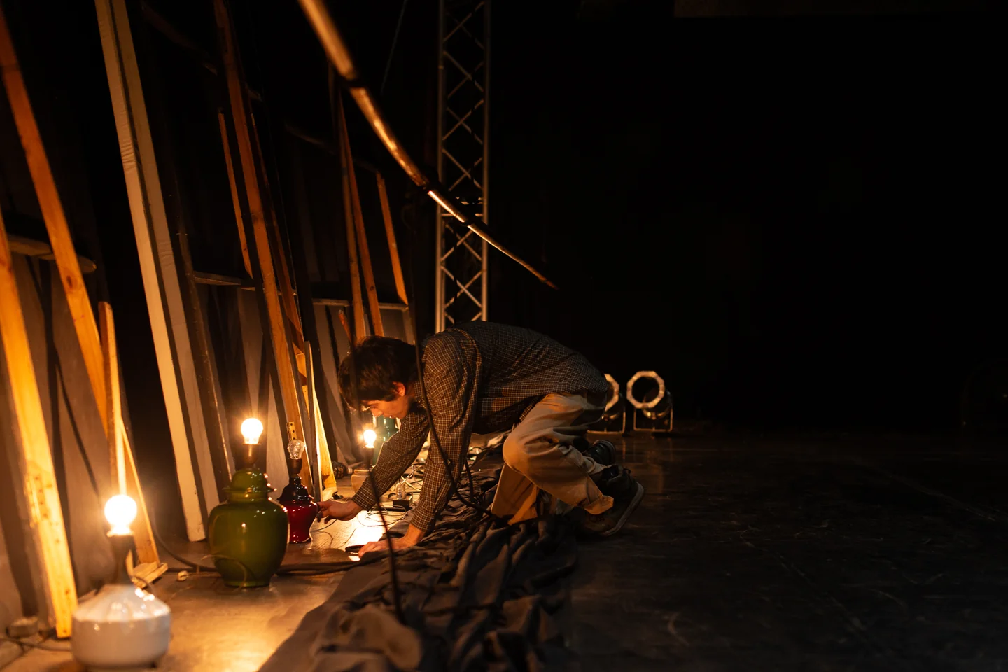 Persona agachada junto a lámparas encendidas, cables y estructuras en un escenario oscuro