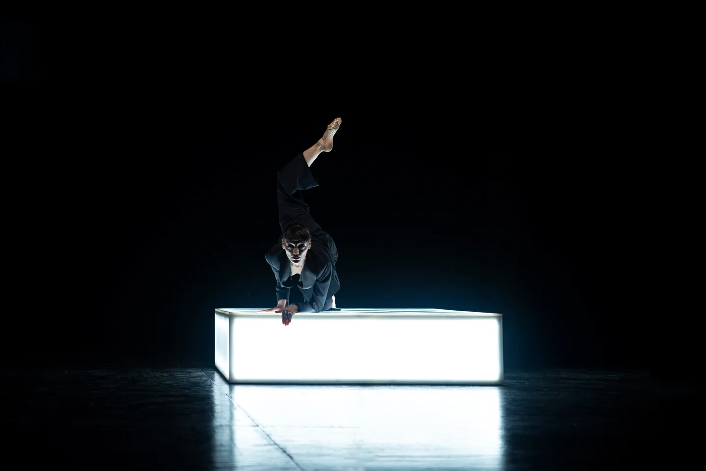 Persona con una pierna elevada y las manos apoyadas en una plataforma luminosa sobre un escenario oscuro