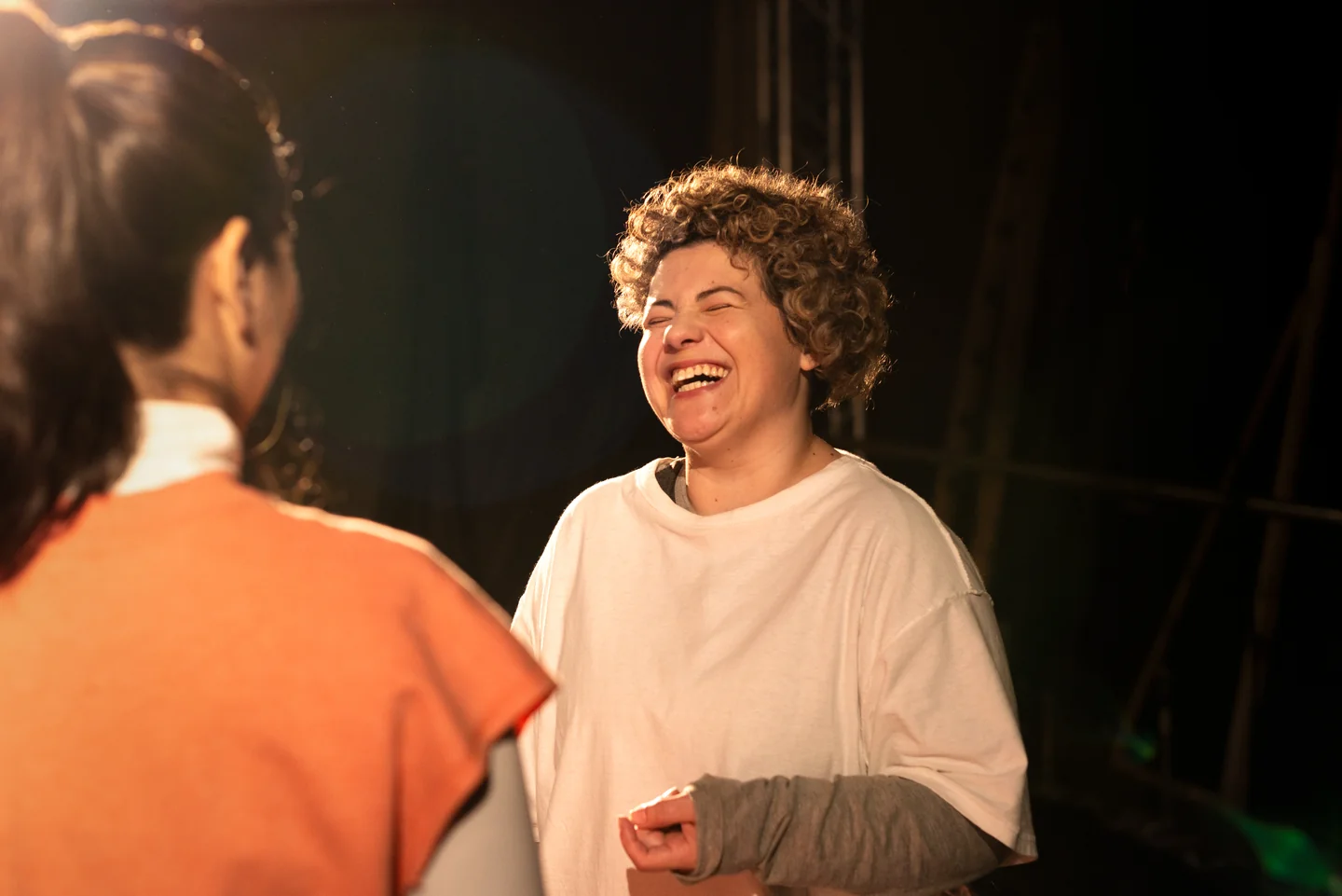 Persona de pelo rizado riendo frente a otra de espaldas con ropa naranja, iluminadas por luz cálida