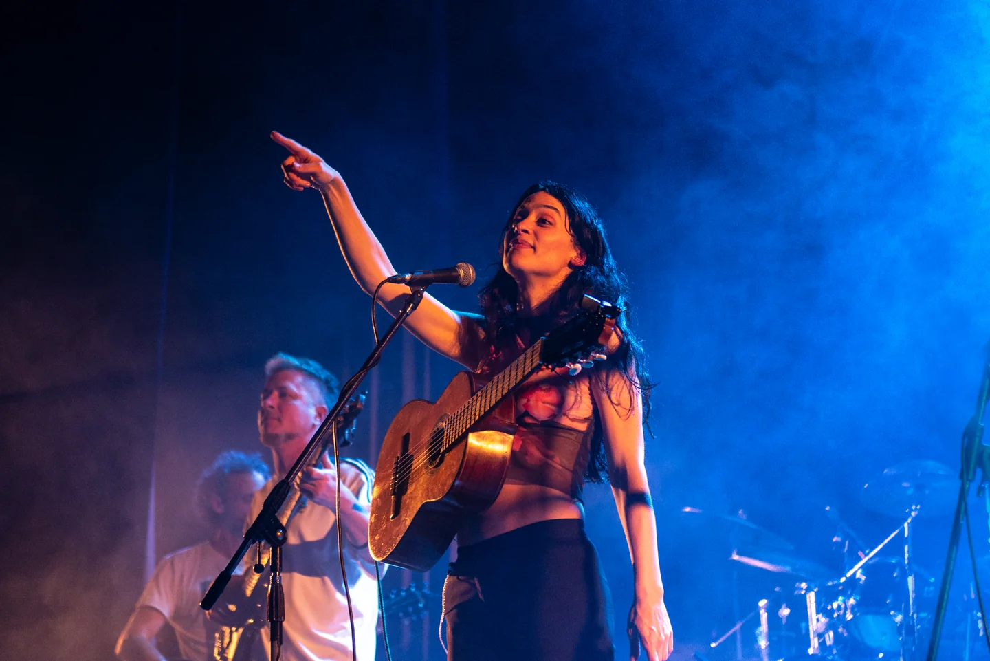 Cantante con guitarra señalando al público junto a un micrófono, bajo iluminación azul