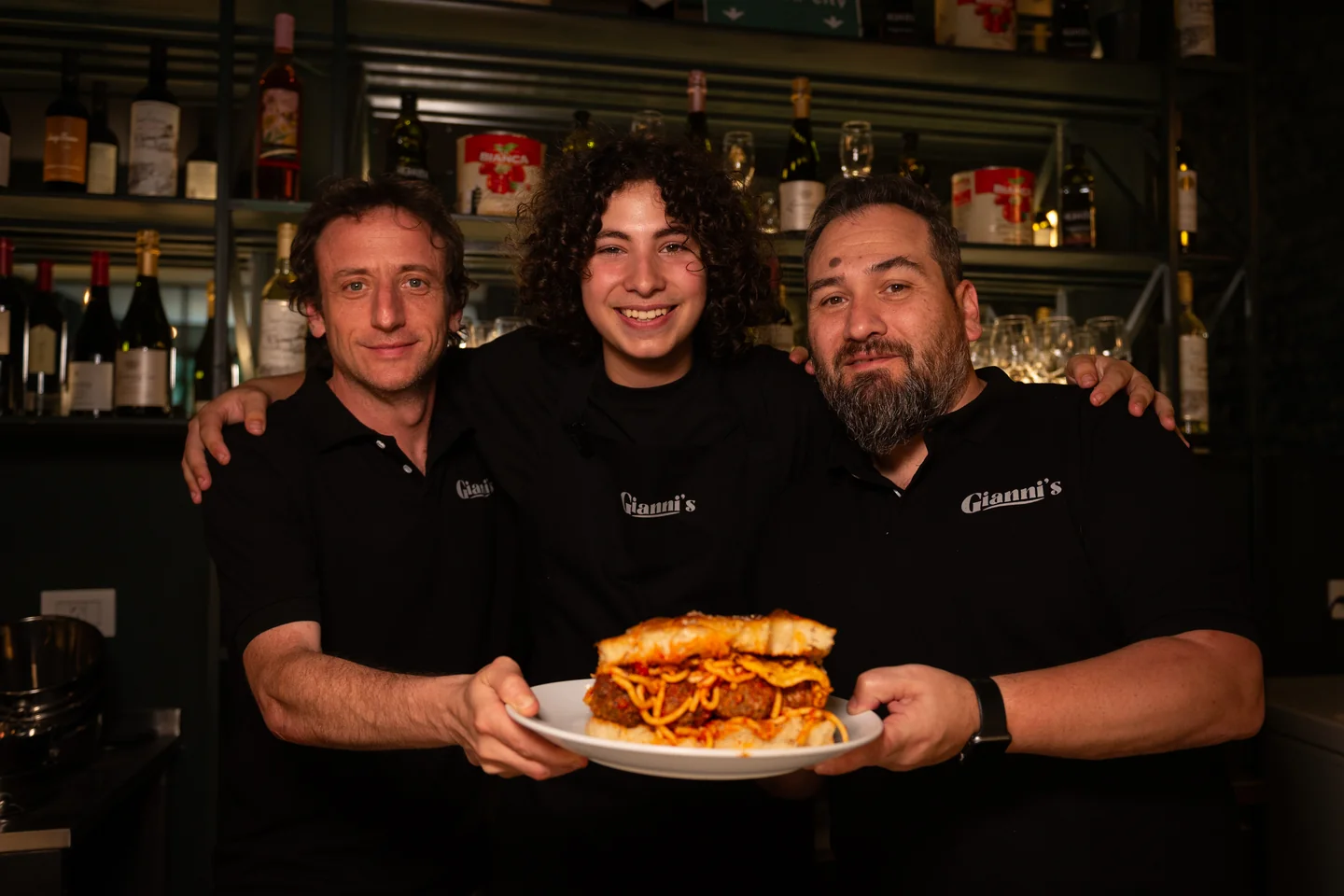 Tres personas sonriendo y sosteniendo un plato ante una barra, con prendas Gianni’s